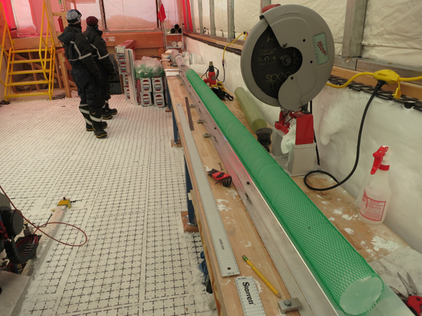 A 1 meter long section of ice core and a 2 meter long section of ice core are matched up on the processing table before the 2 meter long section is cut into two 1 meter long sections using a dry-cut circular saw. The ice shown in this photo is from the 'brittle ice zone'. Because of the potential brittle behavior of this ice, the ice is extruded into (green) plastic netting after it is drilled to ensure that if any breakage of the ice occurs during handling or transport the core fragments will be contained. Photo credit: Murat Aydin